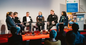 Sieben Personen sitzen im Halbkreis auf dem Podium und diskutieren.