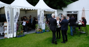 Menschen stehen an Stehtischen und unterhalten sich. Im Hintergrund ist der Infostand zu sehen.