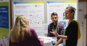 Zwei Frauen und ein Mann stehen um einen Tisch und unterhalten sich. Im Hintergrund sind zwei Poster am Infostand des Monitoringzentrum zu sehen